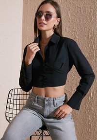 Black cropped shirt with long sleeves and buttons paired with high-waisted light gray jeans. Model wears sunglasses and gold earrings.