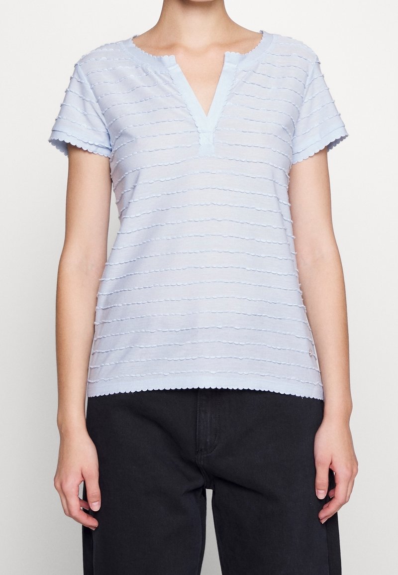 Haut bleu clair à manches courtes avec un décolleté en V, présentant des rayures horizontales texturées et des bords festonnés. Porté avec un pantalon foncé.