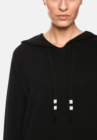 Schwarzer Kapuzenpullover mit V-Ausschnitt, aus weichem Strickmaterial gefertigt. Mit schwarzen Kordeln und weißen Akzenten. Schlichte Textur, lässiges Design.