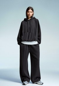 Kvinde står iført en oversized sort hoodie, løse sorte bukser, hvid t-shirt under og grå og hvide sneakers på en ensfarvet lys baggrund.