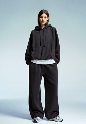 Woman standing wearing an oversized black hoodie, loose black pants, white undershirt, and gray and white sneakers on a plain light background.
