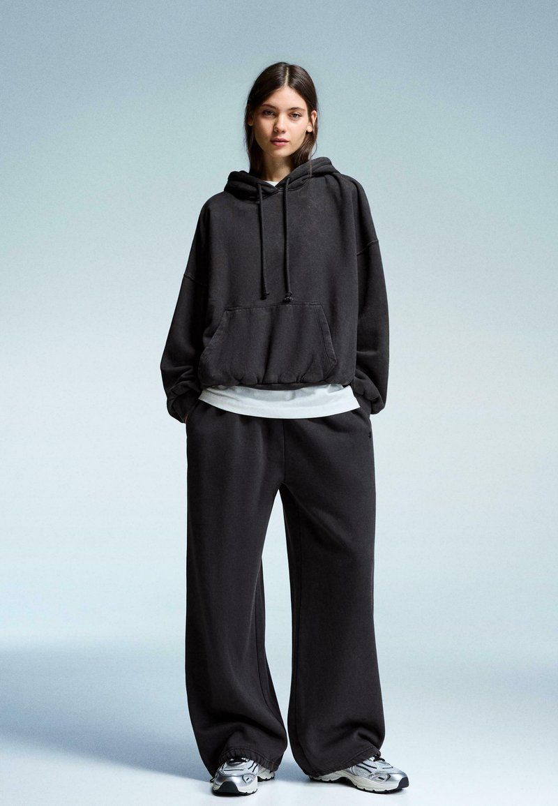 Kvinde står iført en oversized sort hoodie, løse sorte bukser, hvid t-shirt under og grå og hvide sneakers på en ensfarvet lys baggrund.