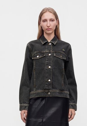 Femme aux cheveux blonds et raides porte une veste en jean noire avec fermeture par boutons et poches devant, sur une jupe noire, face à elle.
