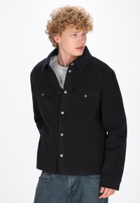 Giacca camicia nera in lana con chiusura a bottoni, due tasche sul petto e bottoni a pressione argentati. Tessuto strutturato con design del colletto classico.