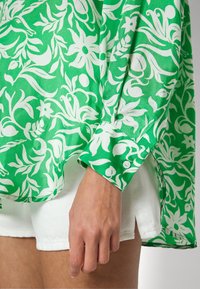 Chemisier vert à motifs avec des dessins floraux blancs, doté de poignets à boutons et de fentes latérales, associé à un short en denim blanc.