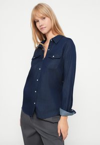 Camisa de ganga azul-marinho com mangas longas, dois bolsos no peito, fecho com botões à frente e punhos de ganga em contraste. Textura suave, design clássico.