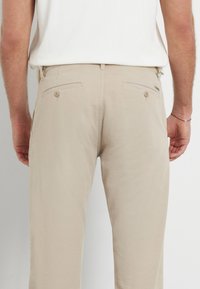 Pantaloni in cotone beige con design a gamba dritta, dotati di due tasche posteriori e chiusura a bottone. Stile minimalista, senza motivi visibili.
