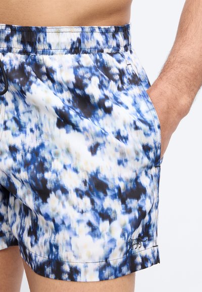 Ο κορμός και τα πάνω μέρη των ποδιών ενός άνδρα που φοράει σορτς με αφηρημένο μοτίβο σε μπλε, λευκό και μαύρο, με το χέρι να ακουμπάει μέσα στην δεξιά τσέπη.