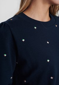 Marinblå sweatshirt med runt halsringning, med spridd metallisk broderi i olika färger, inklusive grön, guld och silver.