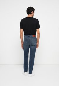 Jeans de denim azul con corte recto, ligero desgaste y bolsillos traseros. Un parche de cuero marrón en la cintura añade detalle.