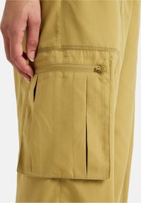 Pantaloni cargo beige con tasca frontale con zip, pieghe sui lati e tessuto liscio con una texture sottile.