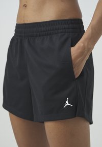 Gros plan de l'abdomen et des cuisses supérieures portant un short de sport noir avec une taille élastique, une poche latérale et un petit logo Jumpman blanc sur la jambe gauche.