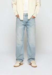 Jeans en denim bleu clair à coupe décontractée, avec une fermeture à boutons sur le devant, cinq poches, et un léger délavage au niveau des jambes. Portés avec des baskets beiges.