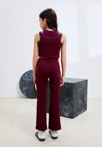 Completino bordeaux composto da top corto senza maniche e pantaloni svasati. Presenta accenti blu e un design aderente. Le sneakers sono bianche e marroni.