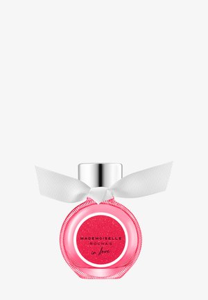 Flacon de parfum rond rose avec bouchon argenté et nœud blanc, étiqueté « Mademoiselle Rochas in Love » sur un fond rouge pailleté.