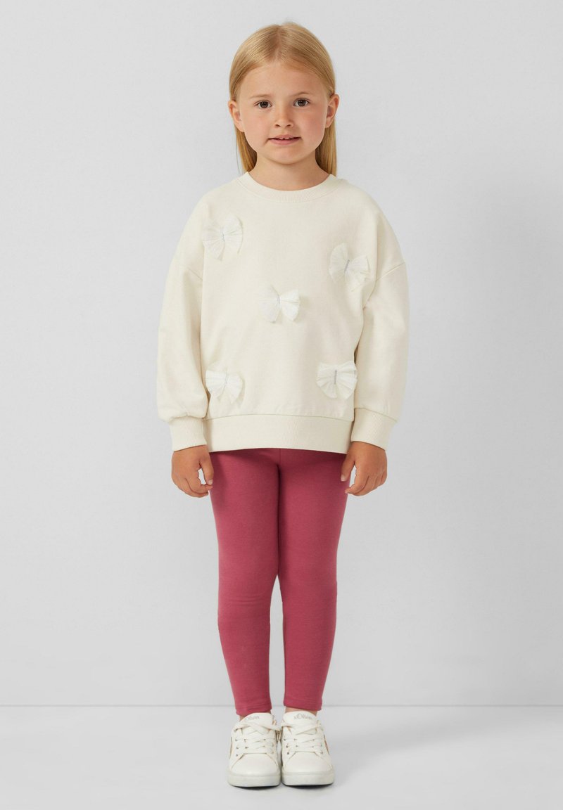 Cremefarbener Sweatshirt mit weißen Stoffschleifen, oversized Schnitt, kombiniert mit pinken Leggings und weißen Sneakern, mit einer glatten Textur.