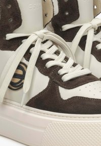 Vysoké tenisky s bílou koženou a hnědou semišovou akcentací, s texturovanou gumovou podrážkou a kontrastními krémovými tkaničkami. Výrazný detail loga.