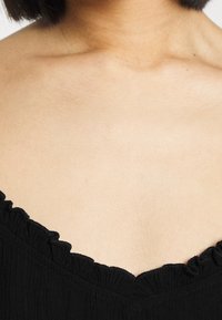 Černý top s volánovým výstřihem, vyrobený z texturovaného materiálu a s hlubokým V výstřihem. Materiál se zdá být lehký a dobře padnoucí.