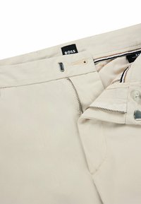 Beige trousers with a zippered front, button closure, and fabric tag labeled "BOSS." Features a smooth texture and stitched accents.