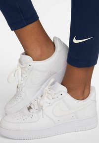 Sneakers bianchi Nike Air Force 1 con punta rotonda, dettagli perforati e lacci piatti; indossati con leggings blu navy con un piccolo logo.