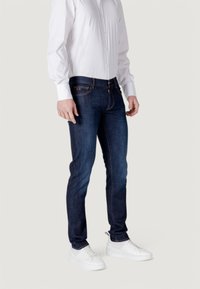 Jeans de mezclilla azul oscuro de corte ajustado, con costuras naranjas y cinco bolsillos. Combinados con una camisa blanca de mangas largas y botones.