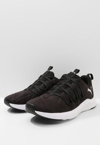 Svarta träningsskor med en vadderad ovandel, texturerad yta, svarta snören och en vit sula. Har en liten logo på sidan.