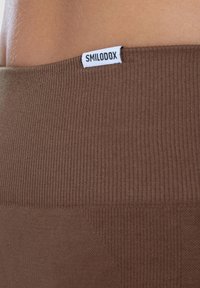 Bruine ribstof met een gladde textuur, met een wit label met de inscriptie "SMILODOX" op de tailleband.