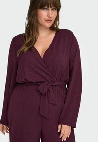 Bordeaux lange mouwen jumpsuit met een v-hals en wikkelontwerp, vastgebonden in de taille. Stof met subtiele glittertjes doorheen. Glad textuur.