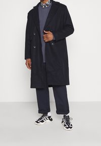 Abrigo de doble botonadura en azul marino oscuro con grandes solapas, llevado sobre una sudadera gris oscuro. Combinado con unos pantalones azul marino enrollados y zapatillas negras.