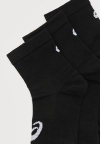 Τρία ζευγάρια μαύρες αθλητικές κάλτσες με λευκή λεπτομέρεια λογότυπου στοιβαγμένες σε λευκή επιφάνεια.