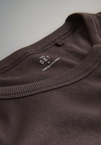 Primer plano del cuello de una camisa de algodón marrón acanalado con una etiqueta impresa que dice "THE SET, M, Made in Bangladesh."