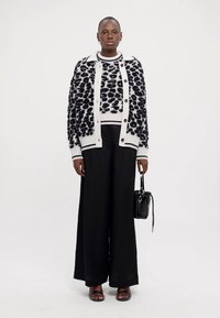 Svart-hvitt mønstret cardigan over en hvit genser, kombinert med svarte vide bukser og svarte sandaler, holder en liten svart veske.