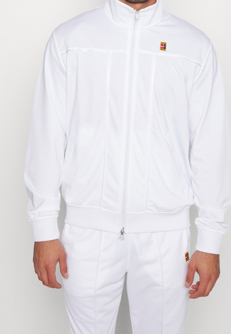 Homme portant une veste de survêtement blanche zippée et un pantalon assorti avec de petits logos carrés rouges et jaunes sur la poitrine et la cuisse.