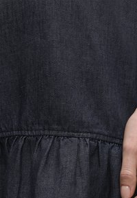 Σκούρο μπλε ύφασμα denim με λεπτομέρεια συρραφής και ένα χέρι με φυσικά νύχια που ακουμπάει στο πλάι.