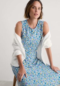 Ziedu raksta bezpiedurkņu kleita gaiši zila, ar violetiem un zaļiem akcentiem. Mīksta auduma, pieguļoša silueta. Valkāta balta kardigāna.