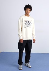 Sudadera crema con estampado gráfico, combinada con pantalones cargo negros que tienen bolsillos laterales y zapatillas plateadas. Tejido suave, ajuste relajado.