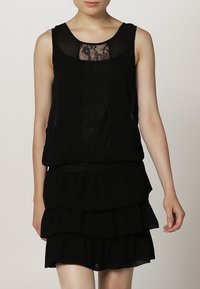 Musta ilmanhiha dressi, jossa on pitsinen yläosa ja kerroksellinen volangihame, pyöreä kaula-aukko ja rento istuvuus. Rakennekangas yksityiskohtana.