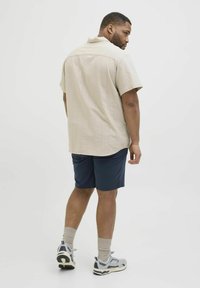 Helles beige Kurzarmhemd mit strukturiertem Muster, kombiniert mit navyblauen Shorts und grauen Sneakern. Sichtbares Rückendesign und Kragen.