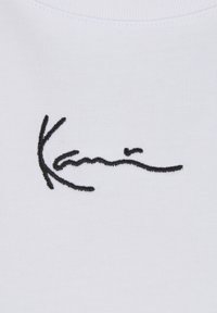 Material alb cu text brodat negru în stil semnătură, cu inscripția „Kamiš” centrată aproape de partea de sus.