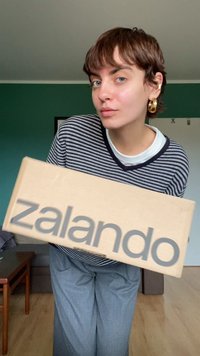 Zalando blagovno znamko kartonska škatla, ki jo drži oseba, v bež barvi, gladki površini in sivo debele črke. Vidna je črtasta majica.