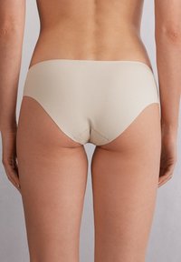 Bežové bezešvé spodní prádlo s nízkým pasem, hladkým povrchem a zakřivenými otvory pro nohy, zobrazené ze zadního pohledu.