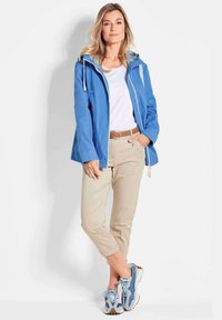 Wasserfester blauerMantel mit Kapuze, beigefarbene Hose und blaue Sneakers. Die Jacke hat einen Frontreißverschluss und verstellbare Kordeln.