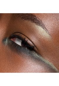 Nahaufnahme eines Auges mit tief schwarzem Augenmake-up und schimmernden grünen Akzenten, einer glatten braunen Hauttextur und gut gepflegten Augenbrauen.