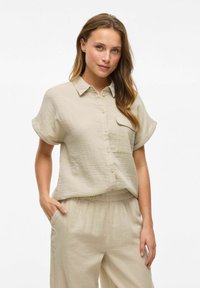 Camicia button-up beige chiaro a maniche corte con colletto e tasca sul petto, realizzata in tessuto testurizzato, indossata con pantaloni larghi coordinati.
