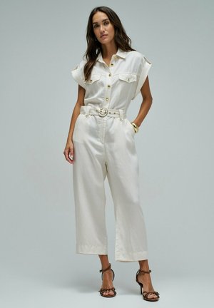 Femme portant une combinaison blanche sans manches ceinturée avec des boutons dorés et des sandales marron à talons hauts, debout devant un fond uni.