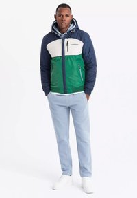 Färgrik zip-jacka med sektioner i marinblått, grönt och vitt; har en huva, två sidofickor och en vit logotyp på bröstet.