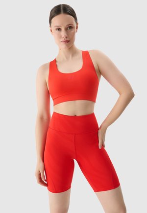 Vrouw met een lichte huid en bruin haar in een rode sportbeha en bijpassende hoge fietsbroek staand tegen een effen lichte achtergrond.