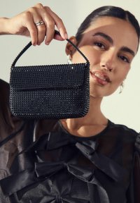 Woman holding black studded handbag by its handle, wearing sheer black top with satin bows and silver hoop earrings.