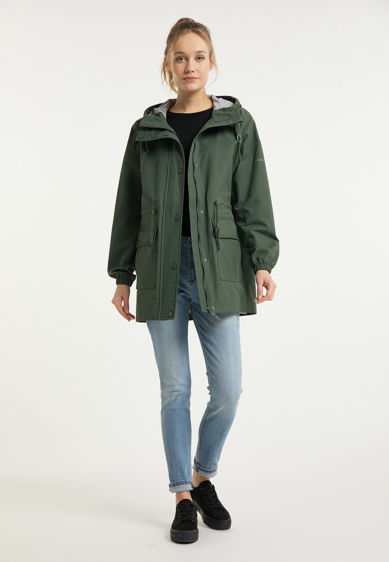 Grüne wasserdichte Jacke mit Kapuze, elastischen Bündchen und zwei Fronttaschen. Kombiniert mit hellblauen Jeans und schwarzen Schuhen.