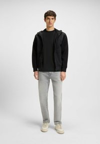 Black hooded jacket with two front pockets over a dark T-shirt, paired with light gray jeans and cream sneakers. Simple, functional design.